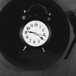 Alarm clock on black plate with cutlery on grey background