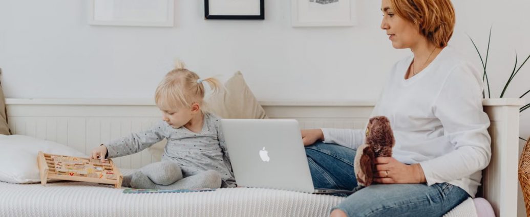 A mother and daughter bonding indoors, sitting on a sofa with a laptop and wooden toy.