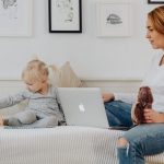 A mother and daughter bonding indoors, sitting on a sofa with a laptop and wooden toy.