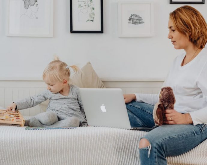 A mother and daughter bonding indoors, sitting on a sofa with a laptop and wooden toy.